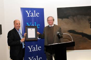 Andreas Dracopoulos, President of the Stavros Niarchos Foundation, holds a glass of champagne and with his other hand he and another man on the podium hold a gift of honor and are photographed in front of a banner that reads Yale